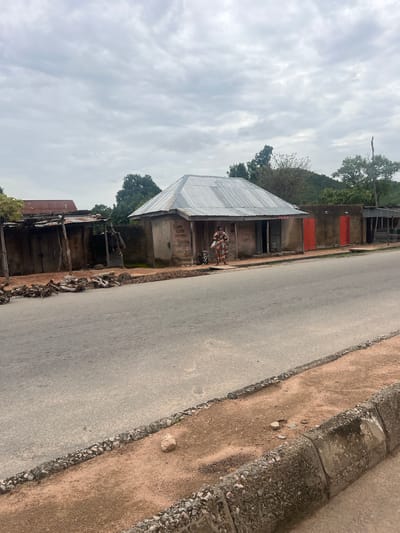 The image depicts a street scene in Kona Garu, Nigeria. A person is standing in front of a building with a corrugated metal roof. The building appears to be a shop or dwelling. Next to it, another structure with a similar roof is visible, and further down the street, more buildings and trees can be seen. The road is paved and runs alongside a sandy embankment with a stone border. The sky is overcast, suggesting cloudy weather. The overall scene appears to be a typical day in a Nigerian town or village. There is some text visible on the wall of the building in the foreground, but it is illegible.