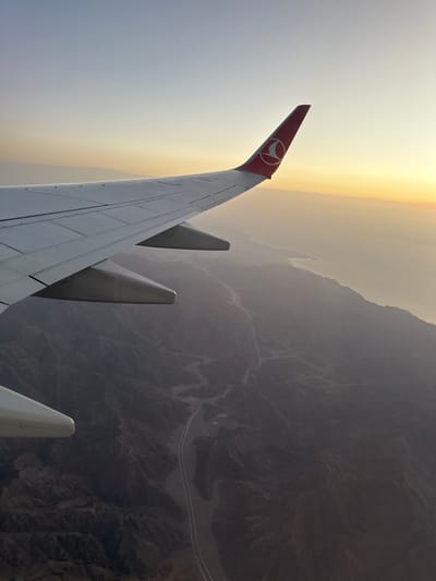 The image shows an aerial view from an airplane, likely taken over a desert region near Dahab, Egypt. The wing of a Turkish Airlines plane is visible in the foreground, marked with the airline's logo on the winglet. Beyond the wing, the landscape appears to be arid and mountainous, with dry riverbeds or valleys carved into the terrain. A road or highway meanders through the landscape. The sky is hazy, with a gradient of light orange or yellow near the horizon, suggesting sunrise or sunset.