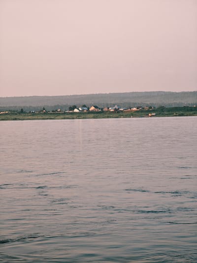 The image depicts a wide river in the foreground, with gentle ripples disturbing its surface. In the mid-ground, a treeline forms a dense barrier of green. Beyond the trees, on a slightly elevated bank, a village is visible. The village consists of several small, simple houses, varying in color from white to natural wood. Some of these structures appear to be residential, while others might be outbuildings or storage units. A few scattered vehicles, possibly trucks or vans, are parked near some of the buildings, suggesting a functional, working village. The background is dominated by a pale, hazy sky, hinting at either dawn or dusk, or perhaps atmospheric conditions like fog or smoke, which softens the distant hills. The overall impression is one of a remote, rural settlement situated along a significant body of water, likely in a forested region. Based on the provided context, this scene is located in the city of Lesosibirsk, Russia.