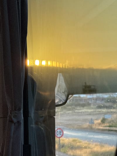 The image shows a view from inside a vehicle, likely a bus, looking out through a window. On the left, a dark gray curtain is drawn, partially obscuring the view. The window is reflecting the bright, golden light of a sunset or sunrise, creating a hazy, atmospheric effect across the top of the image.
Through the window, a landscape is visible. There is a road with a speed limit sign (circular, red-bordered, with "30" inside) on the left. Beyond the road, dry, grassy terrain stretches out, dotted with sparse vegetation and what appear to be distant, low-lying structures. A partial reflection of the vehicle's interior, possibly a rearview mirror or sun visor, is visible in the middle of the window. The overall impression is of a journey through an arid or semi-arid region during twilight. The location is not specified in the image, and therefore cannot be placed in Silifke, Turkey.