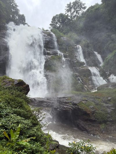 The image shows a multi-tiered waterfall cascading down a rocky cliff face. The water is white and frothy, creating a mist that hangs in the air. Lush green vegetation covers the surrounding rocks and climbs up the cliff face. The sky above is overcast and appears hazy. In the foreground, there is dense foliage with large green leaves and some smaller plants. The scene is located in Ban Sop Aep, Thailand, a region known for its natural beauty and waterfalls. The image depicts a powerful natural display of water and rock, with a serene and somewhat mystical atmosphere due to the mist and the overcast sky. There are no people visible in the image.