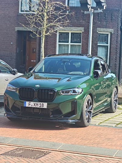 A dark green BMW X4 M Competition, bearing German license plate F XM 18, is parked facing forward on a light-colored paved surface in Wierden, Netherlands. The vehicle features a prominent black kidney grille, an aggressive front bumper with a black lower splitter, and multi-spoke black and polished alloy wheels. No occupants are clearly visible.
Behind the vehicle stands a multi-story brick building with reddish-brown bricks and multiple white-framed windows, some incorporating decorative leaded glass in their upper sections. A wooden front door, partially ajar and displaying the house number 16 to its left, is visible. A deciduous tree with bare branches is positioned in front of the building, between the door and a window. To the right of the tree, a grey utility pole supporting multiple dark grey lighting or utility fixtures extends upwards. The immediate foreground consists of a reddish-brown brick pavement, separated by grey concrete curbs, with a rectangular metal drainage grate visible on the left. A portion of a light-colored vehicle (possibly silver or white) is visible on the far left, parked behind the BMW. The scene is brightly illuminated by sunlight, casting clear shadows.