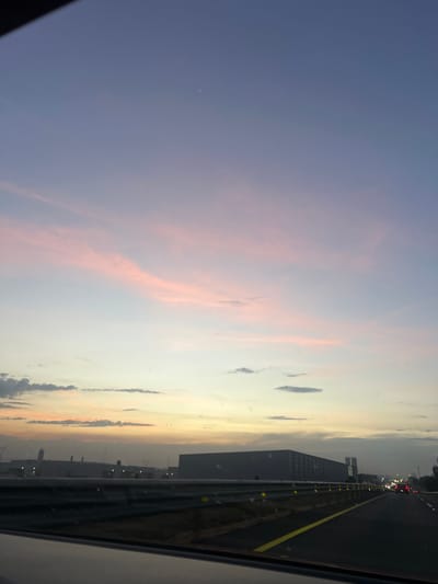 A vibrant sunset paints the sky with hues of pink, orange, and blue. Wispy clouds stretch across the horizon, adding depth to the scene. The view is captured from a car traveling on a highway. A large building sits alongside the road, with other cars visible in the distance. A yellow line marks the road edge, highlighting the perspective of the moving vehicle.