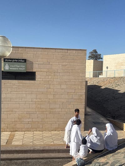 The image captures an outdoor scene at the Miqat Wadi Mahram facility in Al Mashayikh, Saudi Arabia, under a clear, bright blue sky. A building constructed of light-colored, rectangular stone blocks dominates the left and central foreground. A green sign is affixed to the building, displaying Arabic text and "WOMEN'S TOILET" with a corresponding female pictogram. A black rectangular vent is positioned below the sign.

In the foreground, a paved area with light-colored geometric tiles transitions into darker steps. Six individuals are present. Three young males, dressed in white garments, stand on the paved area near the building. One, with dark hair, looks towards the camera. Another stands with his back partially turned, facing towards a group on the right, with a third male partially obscured behind him. To the right of the standing males, three individuals, also clad in white garments and head coverings, are seated on a lower ledge. They appear to be resting or engaging in conversation. A light-colored plastic bag rests on the ground near the seated group.

On the far left, a light-colored, spherical, caged streetlamp fixture is partially visible. In the background, beyond the main building to the right, additional light-colored block structures are visible, along with a sloping terrain exhibiting distinct shadows. A single tree crests the background structures, and a blue railing runs horizontally above the slope. Bright sunlight illuminates the scene, casting sharp shadows.