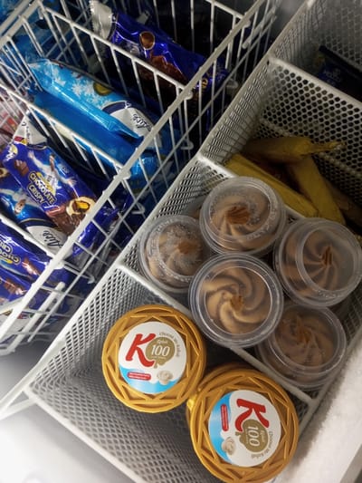 The image depicts the interior of a commercial freezer in Naung Bo Oe, Myanmar, organized with various packaged frozen goods. White wire baskets divide the space. The left side contains multiple individually wrapped ice cream or frozen dessert bars in blue packaging; some are labeled "CHOCOLATE CRISPY" with "Messi" branding, and others bear the "Alice" brand with snowflake patterns. The right side of the freezer holds a white perforated plastic basket. Within this basket, several yellow-wrapped cylindrical items are visible at the top. Below these, a cluster of at least six clear plastic cups, each with a clear lid, contains a swirled brown and lighter-colored frozen dessert, topped with a small central embellishment. In the lower right of this section, two round, golden-yellow containers are present, featuring "K 100 Kaluli" branding and "Choose my Kaluli" text. Frost is visible along the right interior edge of the freezer. No individuals are present, and no actions are occurring.
