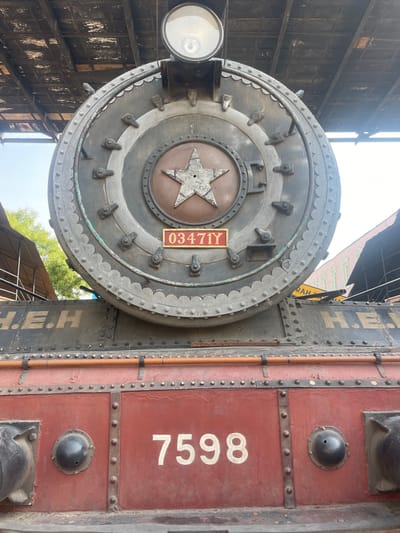 The image shows a close-up of a portion of a vintage steam locomotive. The front face of the boiler is visible, featuring a large circular door with a raised star emblem in the center and the number "0347IY" on a small plate. A headlight is mounted on the left side of the boiler. Adjacent to the boiler is a maroon-colored side panel with the number "7598" painted in white. The environment includes parts of a building with a dark facade. The location is implied to be Anajpur, India.