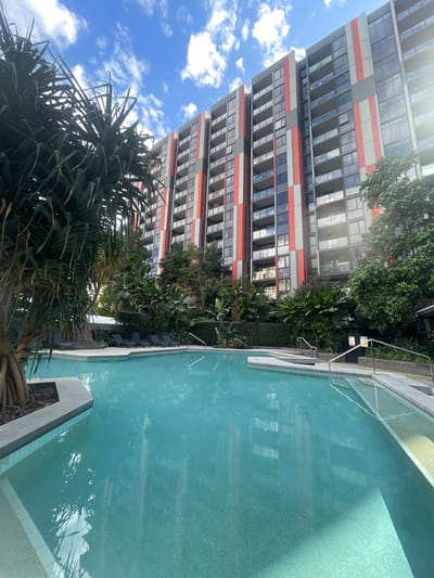 The image shows a swimming pool in front of a modern apartment building in Brisbane, Australia. The building is tall and has a grey facade with red and orange vertical stripes. The pool is a bright turquoise color and has a tiled bottom. There are lounge chairs around the pool, as well as lush greenery. The sky is blue with some white clouds. The overall impression is one of a relaxing and luxurious urban oasis.