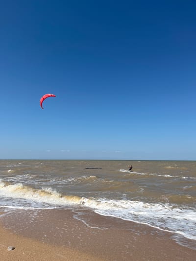 The image depicts a bright, sunny day at a beach. A vast expanse of clear blue sky dominates the upper portion of the frame. In the distance, the brown, choppy water of the sea stretches to the horizon, meeting the sky. A foamy wave is breaking on the sandy shore in the foreground. A person is visible in the sea, actively engaged in kitesurfing, with a red kite high in the sky to the left and the kitesurfer further to the right. The sandy beach is visible at the bottom, with a small, clear object resting on it. The scene appears to be located in Dolzhanskaya, Russia, a location known for its beaches and water sports.