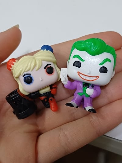 The scene, located in 서대문구, South Korea, depicts two miniature collectible figures, identifiable as Funko Pop! style representations of Harley Quinn and The Joker, resting on the open palm of a human hand. The hand, exhibiting a light skin tone and visible palm lines, occupies the lower and central portion of the frame. On the left, the Harley Quinn figure features blonde hair with prominent red and blue accents, a pale face with black eyes outlined in red and blue, and a red and black costume, holding a black stylized mallet. On the right, The Joker figure is portrayed with vibrant green hair, a pale face, a wide red smile, and is dressed in a purple suit with a green shirt and black shoes; its right arm is raised in a partial waving gesture. The background consists of a uniformly blurred, bright white surface.