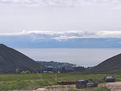 The image depicts a wide landscape scene with a body of water in the background. The foreground consists of a grassy field with some debris, including large pipes and stacked concrete blocks. On the right side of the foreground, a silver sedan with its doors open is parked, and a person wearing a white shirt is standing next to it, appearing to interact with the vehicle.
The middle ground features a town nestled between hills and the water. The town appears to have a mix of residential buildings, some larger structures, and trees. A flag pole with a flag is visible on the left side of the town area. The body of water, likely Lake Issyk-Kul, is calm and reflects the sky. The sky is mostly overcast with some patches of blue visible.
The location is identified as the city of Cholpon Ata, Kyrgyzstan. The scene captures a natural landscape combined with a developed area, suggesting a rural or semi-urban setting. The overall atmosphere is serene, with a touch of everyday life evident by the parked car and the person.