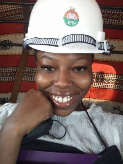 The image displays a frontal portrait of a young adult female located in Lapai, Nigeria. She has dark skin, is smiling broadly with teeth visible, and has prominent false eyelashes. She wears a white hard hat with a black and white striped band, featuring a circular, multi-colored decal on the front depicting a figure, an orange gear-like shape, and a green base. Her attire includes a light-colored, textured top. A black face mask hangs around her neck, resting on her chest. Her right hand is bent at the wrist, with her fist resting near her chin. A purple object, possibly a binder, is partially visible below her hand. The background consists of a patterned fabric or wall covering with horizontal red and beige stripes adorned with black and orange/brown geometric patterns. A dark, vertical wooden element is visible on the left, and a circular highlight of light appears on the right side of the background.