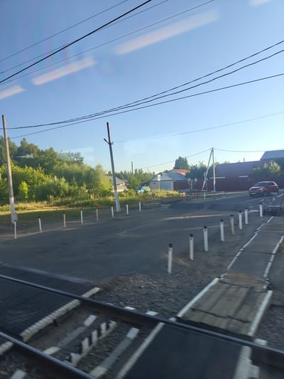 A wide-angle, eye-level, exterior shot captures a view from inside a moving vehicle, looking out at a railway crossing in Isakovo, Russia. The sky above is a clear, pale blue with some faint horizontal reflections on the window. Several power lines crisscross the upper portion of the frame. In the midground, a paved road intersects with railway tracks. Several white posts with black tops line the side of the road. A red car is visible on the road, and further in the distance, there are buildings with red and grey roofs, surrounded by green trees and foliage. The foreground consists of the railway tracks and crossing, appearing slightly blurred due to motion.