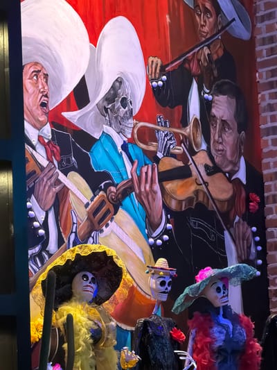 The image displays a vibrant mural of a mariachi band, with the figures depicted as skeletons in traditional charro attire. The mural features individuals playing various instruments, including violins, a trumpet, and guitars. The style of the artwork is colorful and expressive, with bold strokes and a focus on detail in the clothing and facial features of the skeletal musicians. In the foreground, there are three Day of the Dead-themed skeleton figures, dressed in festive clothing and wearing ornate hats. One skeleton is adorned with a large yellow sombrero and a yellow feathered boa, while another wears a green wide-brimmed hat with a pink flower and a red boa. A third skeleton, to the left, wears a straw hat with colorful pom-poms. These foreground figures appear to be decorative or part of a display, with their heads facing forward or slightly to the side. The background of the mural is a solid red color, creating a strong contrast with the figures. The overall scene suggests a celebration or cultural event, likely related to Dia de Muertos or Mexican heritage. This mural is located on the exterior of the restaurant "El Barrio" at 49 Easton Avenue, New Brunswick, New Jersey.