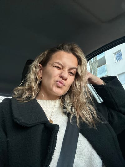 A woman with blonde, wavy hair and a white sweater is wearing a black coat and a seatbelt in what appears to be a car. She is puckering her lips and winking one eye at the camera. An earring is visible in her left ear. Part of a building and sky are seen in the background. The image is rotated.