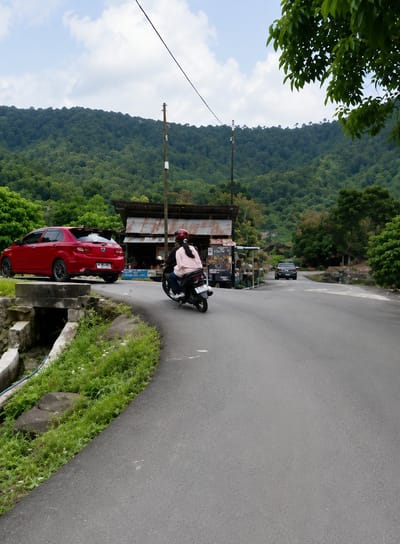 The image depicts a winding asphalt road in Turpuk Sihotang, Indonesia, bordered by lush green vegetation and a steep, tree-covered hillside under a cloudy sky. On the left side of the road, a red hatchback car is parked. Further along, a rudimentary roadside structure with a corrugated metal roof stands beside the road, displaying various items for sale. A man and a woman are riding a scooter, heading away from the viewer. The woman, wearing a pink shirt and a helmet, is driving, while the man, also wearing a helmet, is the passenger. A black car is visible further down the road, approaching from the opposite direction. The overall scene suggests a rural or semi-rural setting in Indonesia.