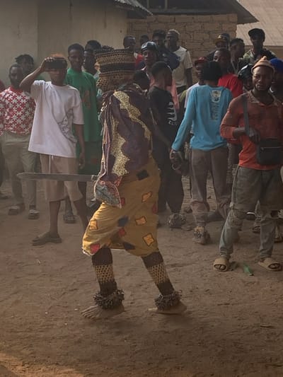 A group of people stands on a dirt ground, some looking at a central figure. The figure is adorned in patterned yellow and purple clothing, with elaborate leg coverings. Many in the crowd are casually dressed, with one person holding up a phone, possibly taking a photo. A building is visible in the background. The scene appears to be an event or performance taking place in an outdoor area.