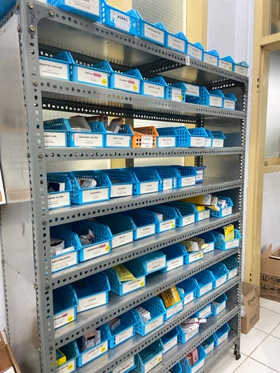 This is a medium-shot, eye-level, indoor photograph of a multi-tiered metal shelving unit filled with blue plastic bins containing various medications. The shelving unit is gray and appears to be a standard pharmacy or medical storage rack. Each blue bin has a white label with black text indicating the medication name and dosage, and some also have yellow "LASA" stickers, likely indicating "Look-Alike, Sound-Alike" medications. The medications are organized alphabetically or by category on the shelves. The background consists of a light-colored wall, a wooden door frame, and a section of a window with vertical blinds. The overall environment suggests a pharmacy, clinic, or hospital storage area. There are no people or animals visible in the image. The specific location of Margoyoso, Indonesia is an external piece of information not derivable from the image itself.