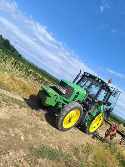 A tilted, eye-level outdoor shot depicts a green John Deere tractor with a red tilling attachment in a rural field under a bright blue sky with white, wispy clouds. The tractor is positioned at an angle, facing towards the right side of the frame. The front of the tractor displays the "6430" model designation and "JOHN DEERE" branding. The tractor has large, black tires with yellow rims. Adjacent to the tractor, on the left, is a field of young green crops, likely corn, with dry, yellowed grass in the foreground. In the background, more rows of green crops stretch towards the horizon, interspersed with trees and distant rolling hills. A power line runs diagonally across the upper left quadrant of the image. The ground appears to be dirt and dry grass. The scene is static, with no people or animals visible. The location is identified as Gamzovo, Bulgaria.