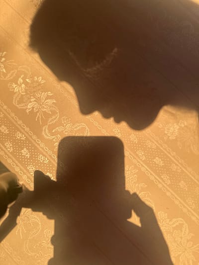 This image depicts an interior scene in Fowlis Easter, United Kingdom. The background is a vertical surface, likely a wall or fabric, displaying a repeating pattern of stylized floral bouquets and geometric bands on a warm, light golden-yellow ground. Strong, warm light from an unseen source, positioned to the upper left, illuminates the surface, creating sharply defined, dark shadows. A prominent shadow of a human profile, facing left, occupies the upper right portion of the image, showing the contours of the forehead, nose, lips, and chin, with textured hair. Centered below this profile, in the lower half of the frame, is the distinct shadow of a rectangular smartphone with rounded corners. To the lower left of the smartphone's shadow, a human hand's shadow is visible, positioned as if grasping the device. The scene captures the interaction of a person holding a smartphone, casting both their silhouette and the device's onto the patterned surface.