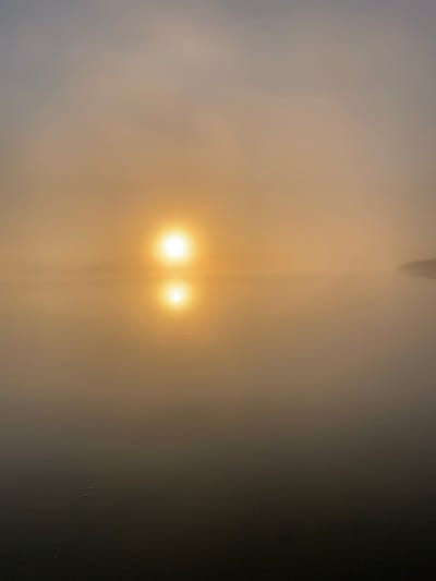 A body of water is shrouded in dense fog, with the sun appearing as a bright, diffused orb near the horizon. The sun's reflection is visible on the water's surface, creating a glowing oval shape. The atmosphere is hazy and muted, with shades of yellow, orange, and gray dominating the scene. The far edge of the water is obscured by the fog, with only a faint suggestion of land visible on the right side. The water itself appears calm and still, with a few small, dark specks dotting its surface. The scene evokes a sense of tranquility and mystery. The location is not specified in the image.