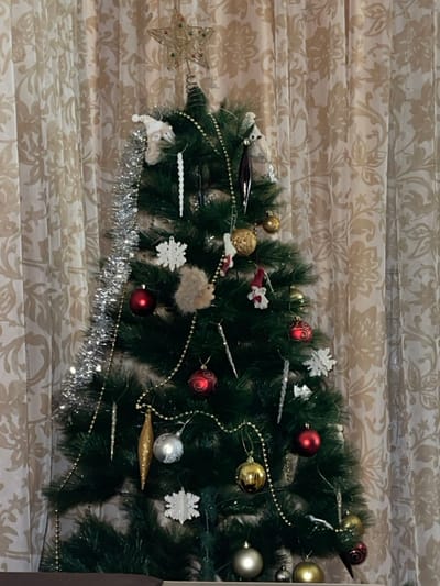 A decorated Christmas tree stands against a patterned beige background. Ornaments include red, gold, and silver balls, snowflakes, icicles, and tinsel. A star sits on the left end. A small, fluffy, round decoration is visible. A string of gold beads is draped around the tree.
