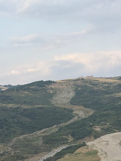 The image displays a landscape view from a high vantage point, looking down a steep, vegetated hillside towards a distant settlement and a road. The scene is dominated by the natural topography, with the hillside covered in a dense growth of green trees and shrubs. The texture of the land is varied, showing rocky outcrops and areas of exposed earth. To the right, a light-colored, gravelly slope descends steeply, with a winding road cutting through it at the bottom.
In the distance, along the crest of the hill, a cluster of buildings is visible, suggesting a village or small town. Some of these buildings appear to have red-tiled roofs. A line of power transmission towers stretches across the landscape, indicating the presence of infrastructure. The sky above is filled with light clouds, suggesting it is daytime, likely late afternoon or early morning given the soft light. The overall impression is of a rural, possibly remote, mountainous region.
There is no visible text in the image. The scene is a static landscape, with no apparent activity or event taking place. The location context of Muğanlı, Azerbaijan, suggests a mountainous or hilly terrain, consistent with the visual information. The image evokes a sense of natural grandeur and the human presence integrated into the landscape.