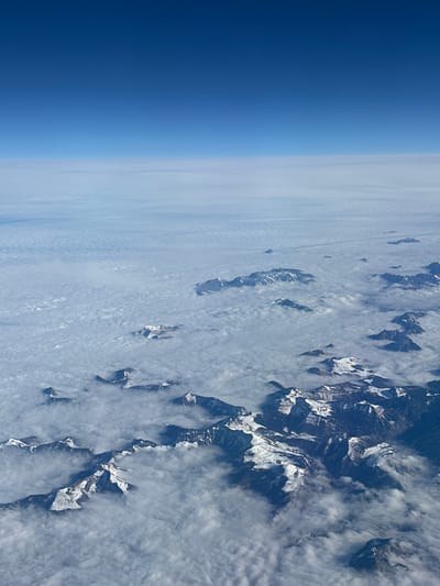 This high-altitude aerial image depicts a vast mountainous landscape within Dimitrovgrad Municipality, Serbia. The upper third of the frame shows a clear, deep blue sky, gradually lightening towards a hazy horizon line in the middle. Below the horizon, an extensive, undulating blanket of white and light grey clouds covers the lower elevations. Numerous rugged mountain peaks, heavily covered in white snow, protrude through this cloud layer. The dark rock formations of the mountain ridges and slopes contrast sharply with the bright snow. No individuals, structures, or any signs of human activity are visible. The scene is illuminated by natural daylight, casting subtle shadows across the snow-covered terrain and cloud formations.