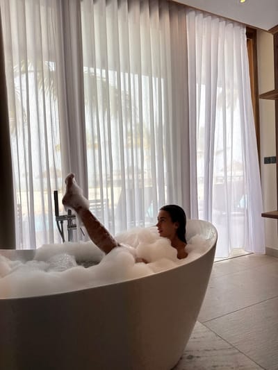 A woman relaxes in a large, white, egg-shaped bathtub filled with bubbly foam. Her dark hair is slicked back, and she gazes downward. Her leg extends out of the tub, her foot visible near the faucet. A bright window covered with white blinds and sheer white curtains is in the background, letting in ample natural light. The bathroom wall to the right appears to have a light gray, textured finish.