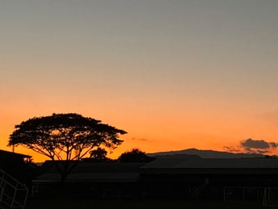 The image displays an outdoor scene at sunset within Chiang Rai City Municipality, Thailand. The sky exhibits a gradient from a vibrant orange-red at the horizon to a lighter orange, transitioning into a blue-grey hue at the top of the frame. In the foreground, a large, deciduous tree with a dense, wide canopy is silhouetted prominently on the left. To the immediate left of the tree, a portion of a white structural element, possibly a frame or railing, is visible. Below the tree and extending to the right, several low-rise buildings with discernible rooflines are silhouetted against the sky. In the dark foreground, which appears to be a field or open ground, two soccer goal frames are visible in silhouette on the right side. In the background, a range of mountains appears as a dark silhouette against the lighter orange sky, with several small, dark cloud formations present above the mountains on the right. No individuals, specific actions, or interactions are observable.