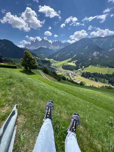 The image shows a first-person perspective of someone sitting on a grassy hillside overlooking the village of St. Magdalena (Santa Maddalena), Italy. In the foreground, the person's legs are visible, clad in light blue jeans and Salomon hiking shoes. To the left, a portion of a metallic object, possibly a fence post or railing, is present. The middle ground features a lush green meadow with scattered white wildflowers, a winding dirt path, and a prominent, mature tree casting a shadow. Beyond the meadow, the village of St. Magdalena is nestled in a valley, characterized by its distinctive church steeple and clustered buildings. The background is dominated by the dramatic, jagged peaks of the Dolomites mountain range under a bright blue sky with scattered white clouds. The overall scene depicts a scenic view from a high vantage point, capturing the natural beauty of the alpine landscape and the charm of the village below.