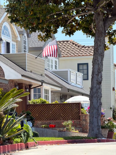 The image shows a residential street in Newport Beach, California. The houses are multi-story, with varying architectural styles and materials, including wood siding, brick, and tile roofs. One house has an American flag displayed. A mature tree is partially visible, obscuring some of the houses. There is a sidewalk and a landscaped area with plants and flowers bordering the street.