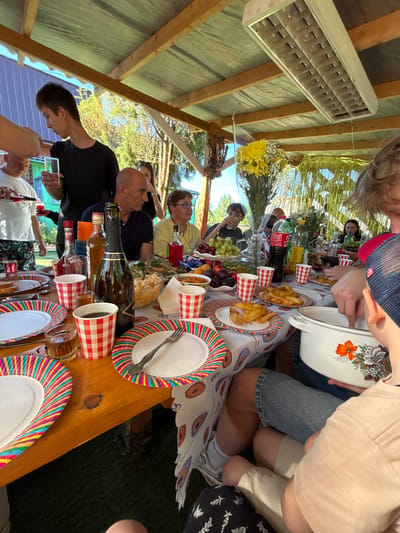 An outdoor daytime gathering is depicted under a covered wooden structure with a corrugated metal roof in Urmanets, Russia. Approximately seven to ten individuals of varying ages, from children to adults, are seated or standing around a wooden table.

The table is covered with a white tablecloth featuring colorful abstract circular patterns. It is extensively set with rainbow-striped paper plates, red and white checkered paper cups, and various dishes. Notable food items include a large platter of green grapes and red/purple plums, plates of fried potato wedges, bowls of what appears to be salad (one with a light-colored chunky salad), and slices of bread. Beverages include a dark bottle of "Puccini Spumante" champagne/sparkling wine, bottles of spirits (one with a red label, another amber-colored), a red ketchup bottle, and bottles of Coca-Cola and a yellow-labeled soda. Several clear glasses containing dark beverages are also present. A prominent bouquet of yellow flowers in clear plastic wrap serves as a centerpiece, with a smaller white floral bouquet further down the table.

In the foreground on the right, a child or young person, wearing a light-colored top and shorts, holds a white enamel pot adorned with an orange and white floral design, with their hand reaching inside. Partially visible next to them is a person with light-colored hair wearing a red baseball cap.

Mid-left, a young male in a black t-shirt stands holding a clear glass. Another male in a white t-shirt with patterned shorts stands to his left, pouring a dark red liquid from a bottle into a glass. Seated at the table are a bald male in a dark blue polo shirt, a female with glasses wearing a yellow shirt, and a young male with glasses in a dark t-shirt who is drinking from a clear glass. Other individuals are partially visible in the background, seated around the table.

The overhead structure consists of unpainted wooden beams supporting a corrugated metal roof, which appears to be light green or blue-green on its underside. A long, rectangular fluorescent light fixture is mounted on the underside of the roof. In the mid-background on the right, golden foil streamers hang as a decoration. The background reveals lush green foliage and trees, and a light blue/purple building wall is discernible in the far left. The ground beneath the table is covered with dark green turf or grass.