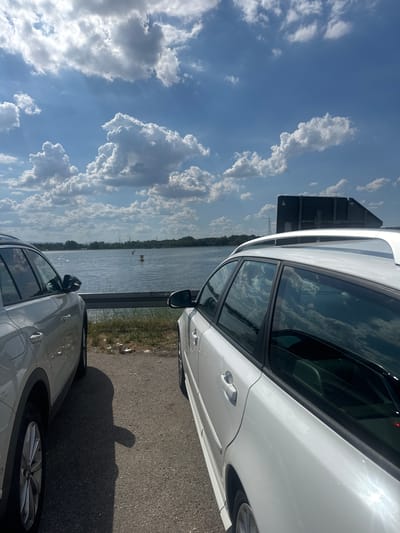 The media file shows a sunlit scene near a body of water, possibly a river or lake, with a backdrop of a bright blue sky filled with dramatic white and grey clouds. Two cars are parked on an asphalt road next to the water. The car in the foreground is a silver SUV, with its front half and wheel visible. The car in the background is a white station wagon, seen from the side, reflecting the sky and clouds in its windows. A shadow of a person, appearing to be making a hand gesture, is cast on the road.
The setting suggests an outdoor recreational area or roadside stop near a natural body of water. The lighting indicates it is daytime, likely mid-morning or afternoon, given the bright sunlight and distinct shadows. The weather appears fair, with abundant clouds but no immediate signs of rain.
The image captures a moment of stillness, with the cars parked and no discernible activity taking place other than the implied presence of the person casting the shadow. The reflections in the car windows add a dynamic element to the scene, mirroring the expansive sky. The overall mood is relaxed and perhaps observational, given the focus on the cars and the surrounding environment. No text is visible in the image.