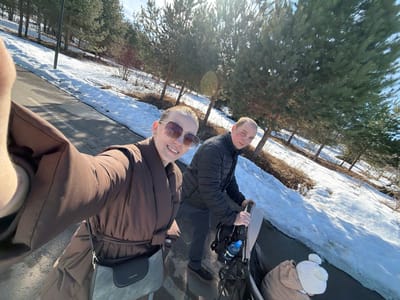 The image shows a family on a paved path in Bezborodovo, Russia, on a sunny day with snow partially covering the ground. In the foreground, a woman wearing a brown belted coat and square-frame sunglasses is taking a selfie, extending her arm to capture the shot, and smiling directly at the camera. A dark grey shoulder bag with a black flap is slung across her body. Behind her, a man wearing a black puffer jacket is pushing a dark-colored baby stroller. A child, dressed in a light brown outfit and a white knitted hat with a pompom, is visible from the back of the head, seated in the stroller. A blue plastic water bottle is attached to the stroller's handle. To the right of the path, the ground is covered in snow, with patches of dry, brown vegetation, and a line of tall evergreen pine trees extends into the background. The path itself is clear and appears wet in places.