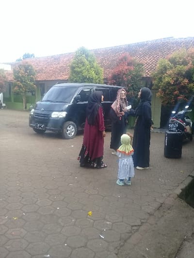 The scene is a street in Kecamatan Cimaung, Indonesia, located at the coordinates -7.068 latitude and 107.554 longitude. There are three women standing in front of a black car. The woman on the left is wearing a black hijab and a black dress, while the woman in the middle is wearing a brown and white dress.  The woman on the right is wearing a red and black dress and a black hijab. A young girl is standing between the two women on the right, facing the camera. The girl is wearing a light green and yellow dress, and her back is to the camera. The woman in the middle is handing the child a gift. The scene appears to be taking place outside a residence with red and brown tile roofing. The ground is paved with grey and white stones. The scene appears to have taken place in the daytime.