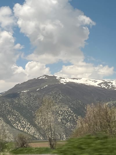 The image shows a mountain partly covered in snow under a sky with scattered clouds. A road and some vegetation are visible in the foreground. The location is not explicitly mentioned in the image.