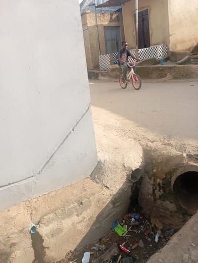 In Gwagwa, Nigeria, a young boy is riding a pink and white bicycle on a dirt road. He is wearing a dark t-shirt and green pants. The boy is positioned in the middle of the frame, with the bicycle angled slightly towards the viewer. To the left of the road, a white concrete wall dominates the foreground. To the right, buildings with checkered patterns and wooden doors are visible. A drainage ditch runs along the edge of the road in the foreground, containing scattered debris and a visible pipe. The overall scene depicts a typical street view in a Nigerian community, with elements of everyday life and infrastructure.