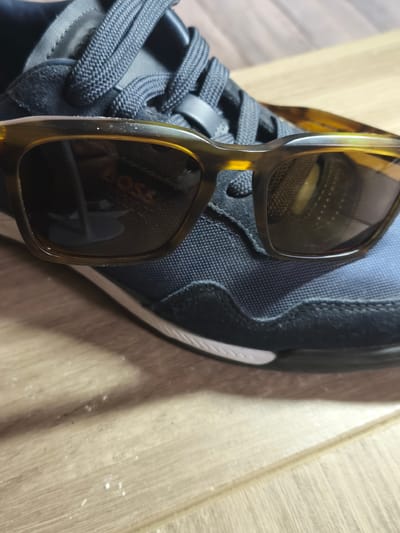 In the city of Fleury-sur-Orne, France, a pair of tortoiseshell sunglasses rests on top of a dark blue sneaker with white laces. The sneaker is made of a combination of suede and mesh materials. The sole of the shoe is partially visible, showing a white stripe. The sunglasses are positioned to partially obscure the laces and the top of the shoe, with the lenses reflecting a blurred image that includes the word "BOSS". The scene is set against a wooden surface, likely a table or floor, which has a light brown, grainy texture and some white dust particles scattered across it. The lighting appears to be artificial and comes from above, casting soft shadows.