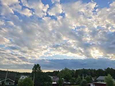 The image shows a sky filled with dramatic clouds, with sunlight breaking through in the upper right quadrant. Below the sky, a line of trees forms a dense forest. In front of the trees, there are several houses and structures, suggesting a rural or suburban setting. The scene appears to be during daylight hours, likely late afternoon or early evening due to the lighting.