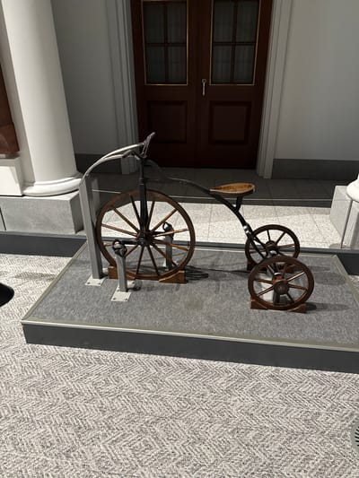 The image depicts a historical bicycle displayed indoors. The bicycle features a large wooden front wheel and smaller wooden rear wheels. It is mounted on a gray display case, which sits on a herringbone-patterned floor. The location is in Sumida, Japan, based on the mention. A dark brown door with gold trim is visible in the background. A white column partially obscures the left side of the bicycle's front wheel.