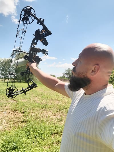 A bald, bearded man in a white t-shirt holds a black compound bow outdoors in a grassy field under a blue sky with scattered clouds. The man is looking up and to the left, with the bow raised. Trees are visible in the background. The scene appears to be located in Semypolky, Ukraine.