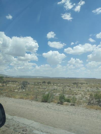 The image depicts a wide, dry landscape under a partly cloudy blue sky, possibly taken from a moving vehicle. The foreground shows a rough, gravelly ground with some cracked concrete or asphalt. A dark rearview mirror is partially visible on the left edge. The middle ground is a vast expanse of dry, sparse vegetation, with scattered bushes and low-lying plants. In the distance, a flat, elevated terrain stretches across the horizon, with some structures or industrial elements visible. The sky is a bright blue, dotted with numerous fluffy white cumulus clouds. The scene suggests a rural or undeveloped area, possibly a desert or steppe environment. The question asks for the location in the city of Besapan, Uzbekistan, however, there is no visual information in the image to support or deny this specific location.