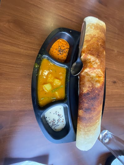An overhead shot shows a close-up of a traditional South Indian meal, likely a dosa, served on a black segmented plate on a wooden table. The dosa, golden brown and rolled, lies to the right of the plate. The plate itself contains three distinct compartments filled with accompaniments. The top compartment holds a vibrant orange chutney with visible seeds. The middle compartment contains a yellow curry with potato chunks and green beans. The bottom compartment features a white chutney, also speckled with seeds. A silver spoon rests on top of the dosa, angled slightly. In the lower right corner, the rim of a clear glass is visible. The overall setting suggests a dining experience in Sidhpur, India, a city known for its rich cultural heritage and culinary traditions.