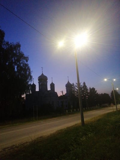The image depicts a street scene at dusk or dawn in Lanivtsi, Ukraine. A prominent street lamp with a bright, starburst-like glare is positioned in the foreground, casting a strong light upwards and outwards. To the left of the frame, silhouetted against the twilight sky, is a large church with multiple domes, the central ones topped with crosses. A large tree with dark foliage partially obscures the left side of the church. A road runs diagonally across the middle ground, with a grassy verge on the right. Several other street lamps are visible further down the road, illuminated. The sky is a deep blue, transitioning to a lighter hue near the horizon, suggesting a time of day when artificial lights are becoming necessary. The environment appears to be a semi-urban or suburban area, given the presence of the church, road, and street lighting. There are no people or animals visible in the scene.