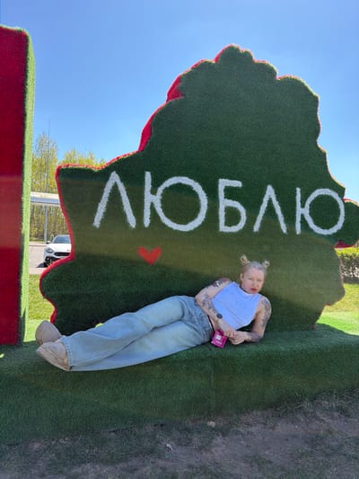 A woman with blonde hair styled in buns is lying on a large, green, map-shaped structure outlined in red, with the Cyrillic word "ЛЮБЛЮ" (meaning "I love" in Russian) spelled out in white. The structure, which appears to be covered in artificial grass, is sitting on a red base. The location is the city of Крошынскі сельскі Савет, Belarus. The woman is wearing a light blue top with the word "Angel" written on it, light blue jeans, and tan boots. She has tattoos on her arms. She is holding a pink object in her hands. The background shows a blue sky and some greenery. A white car is visible in the background.