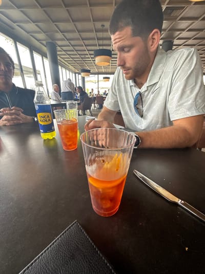 An eye-level, indoor shot shows a man in a light-colored shirt leaning over a table with two orange-colored drinks, a bottle of Inca Kola, and a knife. The taller drink has orange slices inside. Additional people are seated in the background under suspended cylindrical lights. The space appears to be a restaurant with large windows providing natural light. A black leather pad sits on the right side of the table.