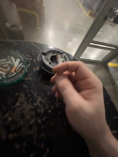 A close-up, slightly out-of-focus image shows a person's hand reaching towards an ashtray filled with cigarette butts. The ashtray is black and circular, with a metallic insert. To the left of the ashtray is a clear glass ashtray also filled with cigarette butts. The surface the ashtrays are on appears to be a dark, possibly stone or laminate table, with a mottled texture. In the background, a reflective surface shows a yellow stripe on a wall and what appears to be a metal structure with glass panels, possibly a partition or railing. The lighting is dim and artificial. The image is taken from a high angle, looking down on the scene. There is no identifiable location in Koziegłowy, Poland.