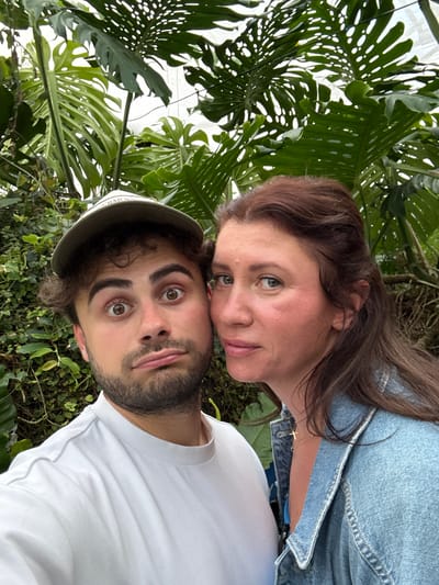Two people, a woman and a man, are posing for a selfie in a lush, green environment. The woman is positioned above and slightly behind the man, with her face close to his. She has brown hair, fair skin, and wears a light blue denim jacket. A small gold cross necklace is visible on her neck. The man has dark hair, a beard, and wears a light-colored baseball cap and a white t-shirt. His eyes are wide and expressive.

The background consists of dense foliage, including large, distinctive Monstera leaves with their characteristic holes. The specific location is not definitively discernible from the image, but it is set in Stratford-on-Avon, United Kingdom.
