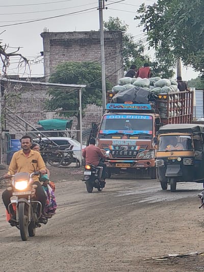 In Walsawangi, India, a street scene unfolds with a convoy of vehicles. A large truck, laden with what appears to be produce wrapped in plastic, is the central focus. Three individuals are seated atop the cargo. Adjacent to the truck is a yellow and black auto-rickshaw with a driver visible inside. In the foreground, a man in a yellow shirt rides a motorcycle, accompanied by a woman behind him. Another motorcycle, with a rider, is seen further down the road. The environment is a dusty, unpaved street with buildings and trees lining the background, under an overcast sky.