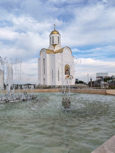 The image captures a view of the Transfiguration Cathedral in Kurgan, Russia, with a prominent fountain in the foreground. The cathedral, a modern white structure with a golden dome topped by a cross, is positioned centrally in the background. In front of the cathedral, a multi-jet fountain sprays water, creating a dynamic visual element. The water in the fountain is a light greenish-blue, and ripples are visible on its surface. The sky above is partly cloudy, with streaks of white clouds against a blue backdrop. Beyond the cathedral, other buildings and structures are visible, including what appears to be a billboard with images and text, and a statue of a figure holding a cross. The overall scene suggests an outdoor public space, likely a plaza or park area, given the presence of the cathedral and the decorative fountain. The time of day appears to be daytime, with soft lighting.